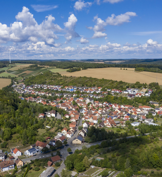 Luftbildaufnahme bei strahlendem Sonnenschein auf den Ort Dittwar. Der Ort liegt inmitten von Feldern und Wald. Im Hintergrund sind Windräder zu sehen. | © Tourist-Info Tauberbischofsheim