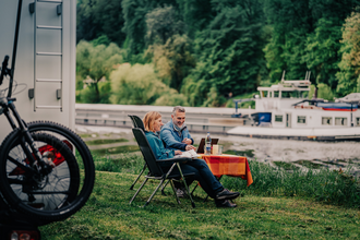 Ein Mann und eine Frau sitzen an einem Campingtisch vor Ihrem Wohnmobil am Ufer eines Flusses. Auf dem Fluß fährt ein Binnenschiff. Auf der anderen Uferseite ist Wald. Neckargemünd im Odenwald. | © Touristikgemeinschaft Odenwald e.V.