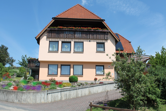 Zweistöckiges Ferienhaus mit roten Dachziegeln, Balkon mit Blumenkästen und gepflegtem Vorgarten unter blauem Himmel | © Ferienwohnung Ehrmann