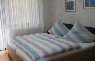 Doppelbett mit gestreifter Bettwäsche in Pastellfarben vor einem Fenster mit weißen Gardinen und einer Wand mit Kreismuster und zwei gerahmten Bildern | © Ferienwohnung Ehrmann