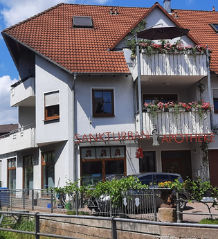 Mehrstöckiges Wohn- und Geschäftshaus mit rotem Ziegeldach und Balkonen mit Blumen, darunter die Sankt Urban Apotheke | © Ferienwohnung Roth