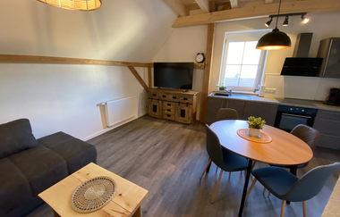 Wohn- und Esszimmer mit grauem Sofa, Holztisch mit vier Stühlen, TV auf Holzkommode und Küchenzeile unter Dachschräge | © Ferienwohnung Etzl