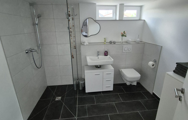 Badezimmer mit begehbarer Dusche, Waschbecken und Toilette, Fließen am Boden schwarz, die Wände weiß | © Wolf