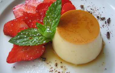 Vanillepudding mit Karamellsoße, garniert mit frischen Erdbeerscheiben, Minzblatt und Puderzucker auf weißem Teller | © Hotel Alexa