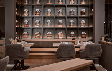 Ein Regal mit beleuchtenden Einmachgläsern. Davor sind drei Tische und Stühle | © Dominik Cini