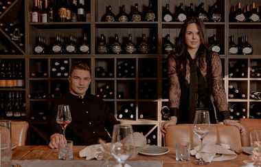 Ein Regal voller Weinflaschen, davor ein Pärchen mit Weingläsern in der Hand | © Dominik Cini