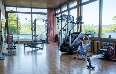Kleiner Fitnessraum mit Hanteln, Kraftstation, Stepper, Heimtrainer und Rudergerät vor großen Fenstern mit Blick ins Grüne | © Vitalhotel König
