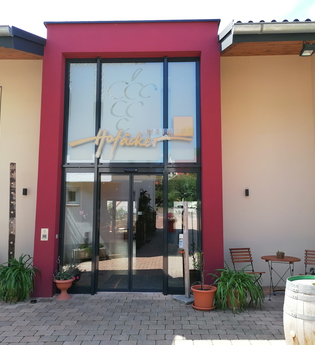 Blick auf den Eingangsbereich mit Glasfront vom Weingut Hofäcker in Weikersheim-Queckbronn. Im Hof davor ist ein Sitzgruppe sowie Blumen und Weinfässer als Dekoration zu sehen. | © Touristinfo Weikersheim