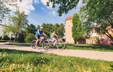 Zwei Radfahrer auf einem Radweg. Im Hintergrund ist eine Stadtmauer mit einem Turm. Eberbach im Odenwald. | © Touristikgemeinschaft Odenwald e.V.