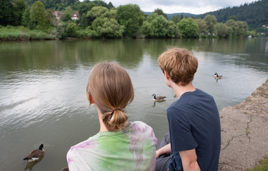 Ein Fluss  mit 3 Enten. An der Ufermauer sieht man eine Frau und ein Mann sitzen und auf den Fluss schauen
