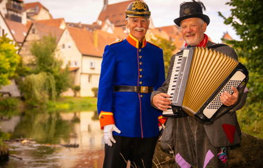 Zwei Männer in historischen Kostümen, einer in einer blauen Uniform und der andere mit einem Akkordeon, stehen vor einem Fluss mit Fachwerkhäusern im Hintergrund. | © Stadt Besigheim