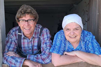 Zwei Personen lehnen sich lächelnd über eine Holztür, eine trägt ein kariertes Hemd, die andere ein blaues Kopftuch. | © SBEntertainment GmbH & Co. KG