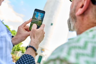 Zwei Herren mit einer Handykamera im Vordergrund, die gerade dabei sind ein Foto vom Hockenheimer Wasserturm zu machen. Auf der Handykamera ist das Gesamtbild des Wasserturms zu sehen. Auf dem Foto erscheint der Wasserturm im Hintergrund. | © Tourimia Tourismus GmbH