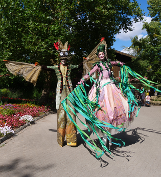 Zwei Personen auf Stelzen in fantasievollen Kostümen in einem blühenden Garten. | © Erlebnispark Tripsdrill
