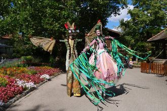 Zwei Personen auf Stelzen in fantasievollen Kostümen in einem blühenden Garten. | © Erlebnispark Tripsdrill