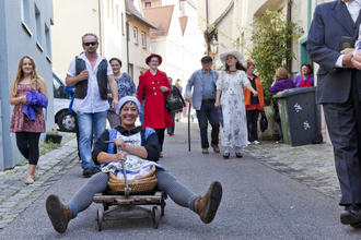 Eine Frau sitzt lachend auf einem kleinen Wagen in einer schmalen Gasse, umgeben von einer Gruppe fröhlicher Menschen. | © Stadt Besigheim