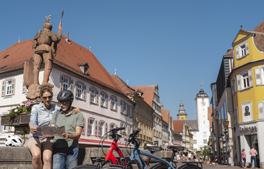 Zwei Fahrräder stehen neben einem Brunnen in der Innenstadt. Die beiden Radfahrer sitzen nebendran auf dem Brunnen und schauen auf einen Stadtplan. | © Philipp Reinhard