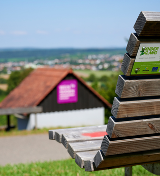 Holzbank mit Rückenlehne im Vordergrund, darauf ein Aufkleber "KinderErlebnisWeg", im Hintergrund ein weiter Ausblick über die Hügellandschaft und ein kleines Häuschen mit lila Infotafel – aufgenommen bei sommerlichem Wetter in Oberderdingen. | © Kraichgau-Stromberg Tourismus e.V.