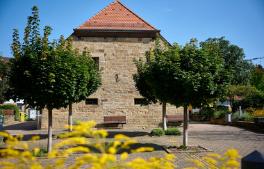 Massives Natursteingebäude mit rotem Satteldach, davor zwei geschnittene Bäume und Sitzbänke – aufgenommen an einem sonnigen Sommertag in Oberderdingen. | © Kraichgau-Stromberg Tourismus e.V.
