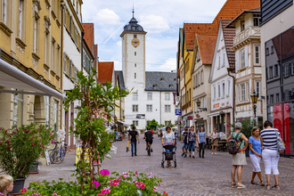 Blick in die Fußgängerzone in der Innenstadt von Bad Mergentheim. Es  sind viele Menschen unterwegs. Im Hintergrund ist das Residenzschloss und im Vordergrund Blumenkübel mit Sommerbepflanzung zu sehen.