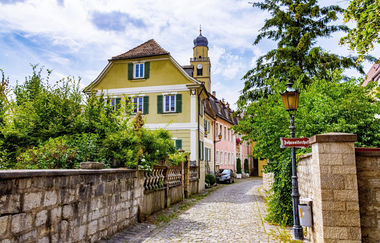 Blick auf ein altes Haus in Bad Mergentheim. Im Hintergrund ist eine Kirche zu sehen. Im Vordergrund eine alte Mauer. Rechts und links des Weges wachsen Büsche und Bäume.