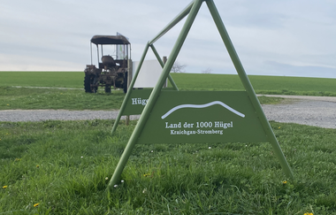 Ein grüner Radständer mit dem Logo „Land der 1000 Hügel“ lädt Gravelbiker*innen zur Pause ein – im Hintergrund ein alter Traktor inmitten der sanften Hügellandschaft. | © Kraichgau-Stromberg Tourismus e.V.