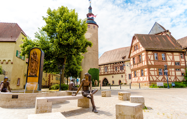 Blick auf den Schlossplatz in Tauberbischofsheim. Im Hintergrund ist der Türmersturm mit dem Kurmainzischen Schloss zu sehen. Im Vordergrund ein Brunnen mit Figuren aus Kupfer. | © Tourismusverband "Liebliches Taubertal" e.V.