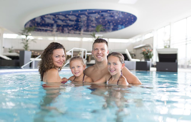 Eine Familie mit zwei jungen Mädchen schaut in die Kamera und lächelt. Sie stehen im Wasser der Innenbecken. | © Solymar Therme