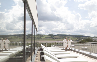 Auf der Dachterrasse befindet sich der Ruhebereich der Saunalandschaft. Bequeme Liegen laden zum Ausruhe ein, eine Frau und eine Mann stehen weiter hinten in Bademänteln. | © Solymar Therme
