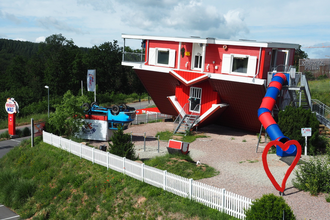 Blick auf das Toppels-Haus "Verdrehte Welt". Das Haus und ein Auto davor stehen auf  dem Kopf. Im Außenbereich ist ein Spielplatz mit Spielgeräten, z.T. auch auf dem Kopf stehend, zu sehen. | © TOPPELS - Verdrehte Welt