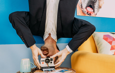 Blick auf einen Mann, der vermeintlich mit dem Kopf nach unten auf einem Tisch steht. Er hat einen Fotoapparat in seinen Händen. | © TOPPELS - Verdrehte Welt