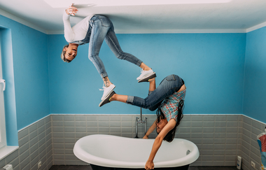 Blick auf zwei Frauen über einer Badewanne. Die eine hängt vermeintlich mit dem Rücken an der Decke, die andere hält sich mit den Händen an der Badewanne fest und der Rest des Körpers zeigt Richtung Decke. Die Füße der beiden berühren sich. | © TOPPELS - Verdrehte Welt