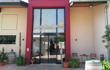 Blick auf den Eingangsbereich mit Glasfront vom Weingut Hofäcker in Weikersheim-Queckbronn. Im Hof davor ist ein Sitzgruppe sowie Blumen und Weinfässer als Dekoration zu sehen. | © Touristinfo Weikersheim