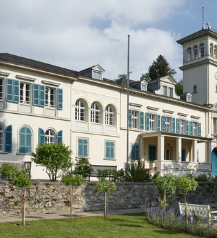 Ansicht des Schlosses mit weißem Verputz und türkisfarbenen Fensterlädem. Zu sehen ist ein quadratischer Turm und ein Teil der Parkanlage. | © Gemeinde Seeheim Jugenheim