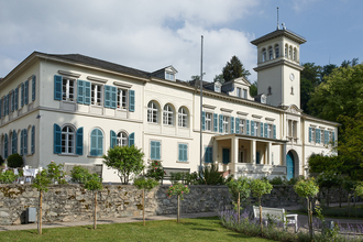 Ansicht des Schlosses mit weißem Verputz und türkisfarbenen Fensterlädem. Zu sehen ist ein quadratischer Turm und ein Teil der Parkanlage. | © Gemeinde Seeheim Jugenheim