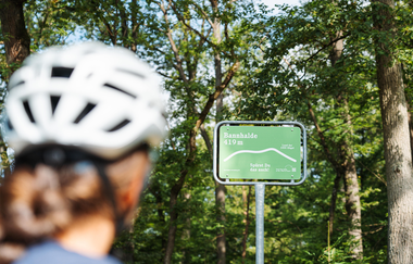 Eine Radfahrerin mit Helm blickt auf ein grünes Schild an der Bannhalde (419 m) im Naturpark Stromberg-Heuchelberg mit der Aufschrift „Spürst Du das auch?“. | © SaddleStories.at