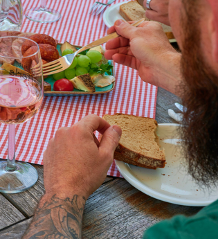 Ein Mann mit Bart und Tattoos genießt eine deftige Vesper mit Wurst, Käse, Trauben und Brot bei einem Glas Roséwein auf kariertem Tischtuch. | © Kraichgau-Stromberg Tourismus e.V.