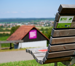 Holzbank mit Rückenlehne im Vordergrund, darauf ein Aufkleber "KinderErlebnisWeg", im Hintergrund ein weiter Ausblick über die Hügellandschaft und ein kleines Häuschen mit lila Infotafel – aufgenommen bei sommerlichem Wetter in Oberderdingen. | © Kraichgau-Stromberg Tourismus e.V.