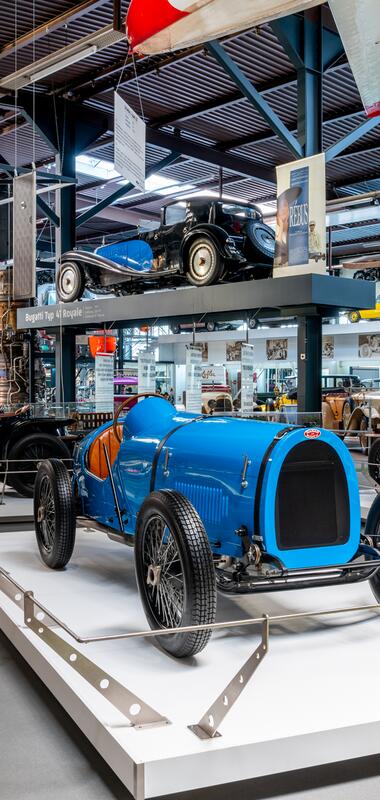 Zwei blaue Bugatti-Rennwagen aus den Anfängen des Automobils, ausgestellt in einem Museum mit weiteren historischen Fahrzeugen und Flugzeugteilen im Hintergrund. | © Technik Museum Sinsheim