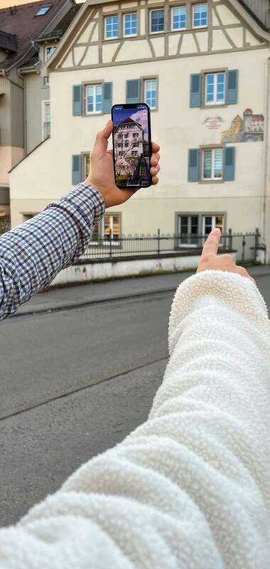 Zwei Personen zeigen mit ausgestrecktem Arm auf ein historisches Fachwerkhaus, das auf einem Smartphone-Bildschirm in einer Augmented-Reality-Ansicht zu sehen ist. | © Foto zeigmal GmbH