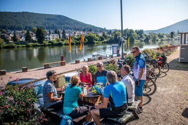 Gruppe von Radfahrern in Sportkleidung sitzt an einem Holztisch am Neckarlauer in Neckargemünd mit Fahrrädern im Hintergrund und Blick auf den Fluss und bewaldete Hügel. | © Landratsamt Rhein-Neckar-Kreis; Foto Adrain Weng