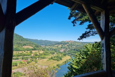 Blick von der Bockfelsenhütte am Neckarsteig durch eine hölzerne Rahmenkonstruktion auf eine Flusslandschaft mit bewaldeten Hügeln und Feldern. Im Hintergrund ist der Dilsberg auf einem Hügel zu erkennen. Der Himmel ist wolkenlos und blau. | © Landratsamt Rhein-Neckar-Kreis; Foto Beate Otto