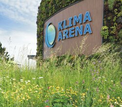 Holzfassade mit dem blauen Schriftzug 'KLIMA ARENA' und einem stilisierten Logo, davor eine Wiese mit Wildblumen und Mohnblumen. | © Landratsamt Rhein-Neckar-Kreis; Foto Dorothea Burkhardt