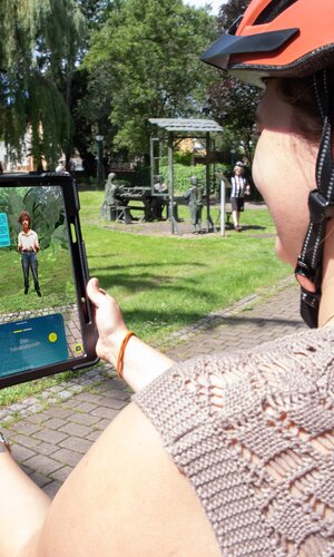 Frau mit orangefarbenem Helm hält Tablet, auf dem eine digitale Figur der Weitersehen App in einem Park angezeigt wird. | © Landratsamt Rhein-Neckar-Kreis; Foto Dorothea Burkhardt