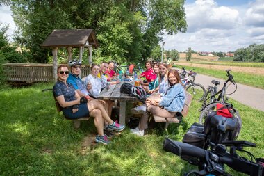 Gruppe von Radfahrern sitzt an einem Holztisch im Freien neben ihren Fahrrädern auf einer Wiese mit Bäumen und einem kleinen Holzdach im Hintergrund | © Landratsamt Rhein-Neckar-Kreis; Foto Dorothea Burkhardt
