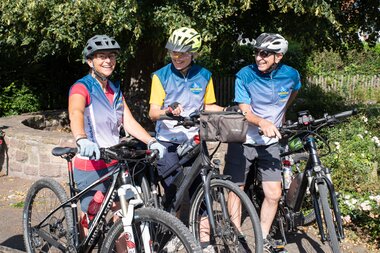 Drei Personen in RadGuides-Trikots mit Helmen stehen mit ihren Fahrrädern vor einer Steinmauer und Büschen. | © Landratsamt Rhein-Neckar-Kreis; Foto Dorothea Burkhardt