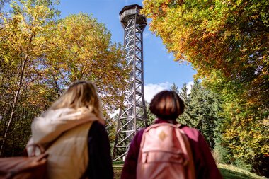 Zwei Personen mit Rucksäcken blicken auf den Teltschikturm, der von herbstlich gefärbten Bäumen umgeben ist | © Landratsamt Rhein-Neckar-Kreis; Foto Sebastian Weindel