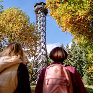 Zwei Personen mit Rucksäcken blicken auf den Teltschikturm, der von herbstlich gefärbten Bäumen umgeben ist | © Landratsamt Rhein-Neckar-Kreis; Foto Sebastian Weindel