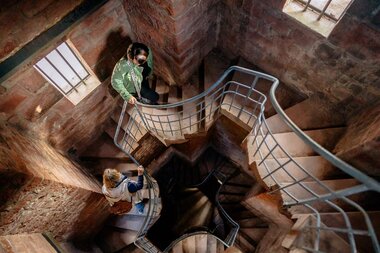 Zwei Personen gehen eine Wendeltreppe mit Metallgeländer in einem engen, gemauerten Treppenschacht hinab. Tageslicht fällt durch vergitterte Fenster auf die Szene. Die Wände bestehen aus rötlichem Stein. | © Landratsamt Rhein-Neckar-Kreis; Foto Sebastian Weindel
