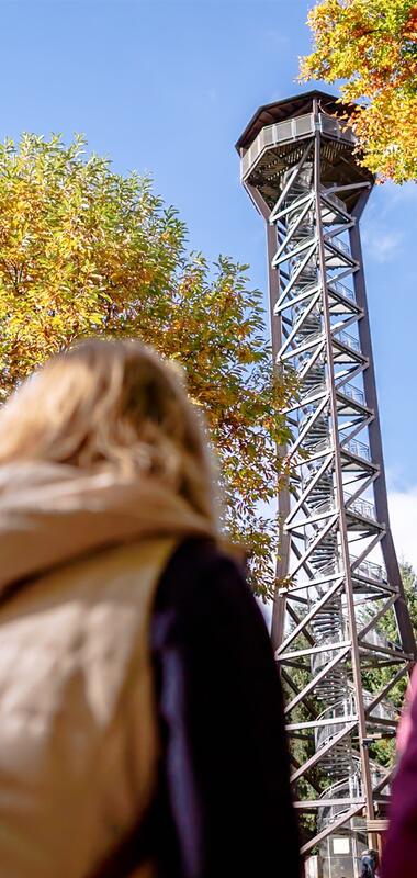 Zwei Personen mit Rucksäcken blicken auf einen hohen Aussichtsturm aus Metall, umgeben von herbstlich gefärbten Bäumen. | © Landratsamt Rhein-Neckar-Kreis, Sebastian Weindel